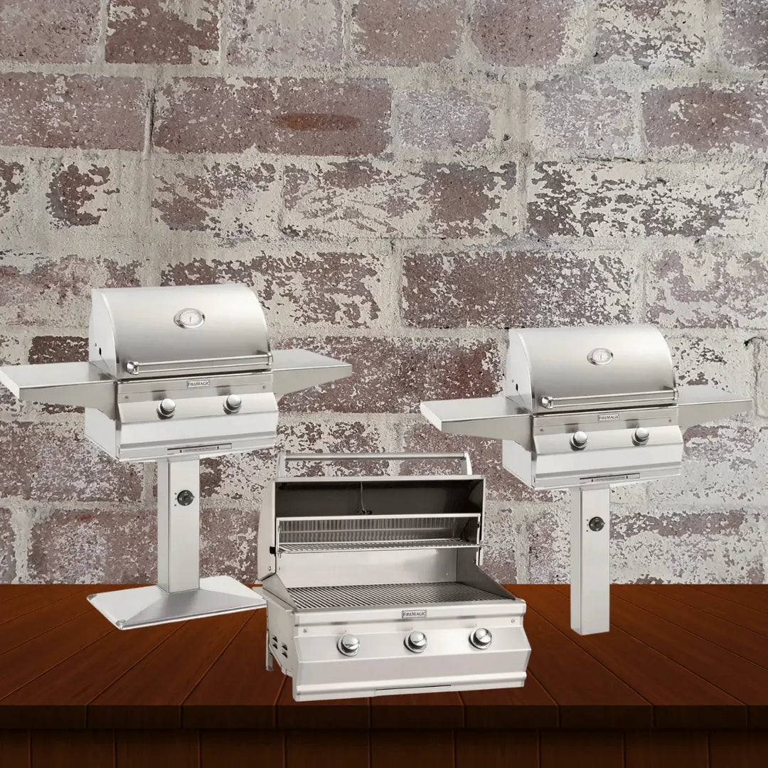 Fire Magic Choice Series gas grills collection featuring compact stainless steel grills, including pedestal-mounted and built-in models with simple control knobs, displayed on a wooden surface with a rustic brick wall background.