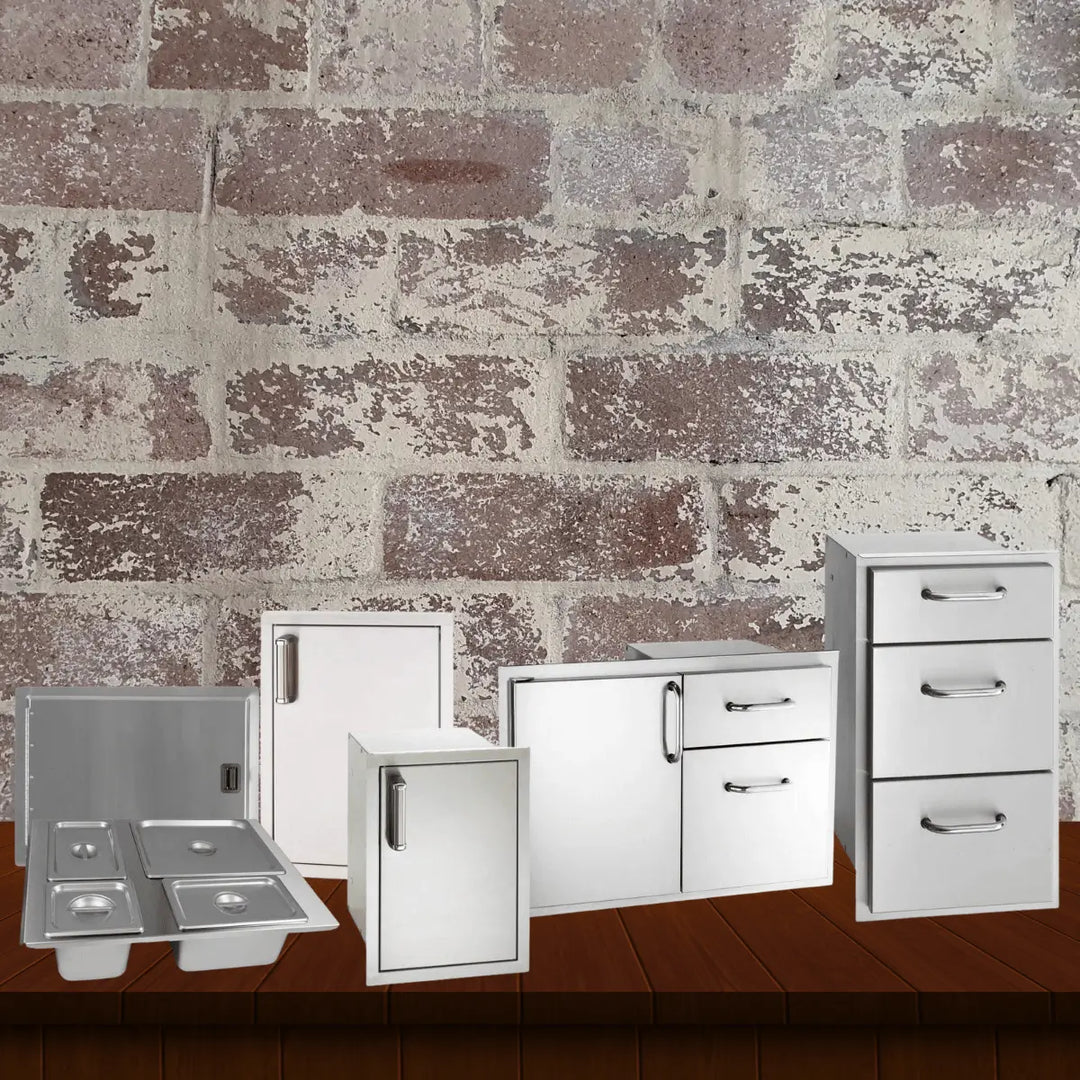 Fire Magic doors and drawers collection featuring stainless steel outdoor kitchen storage components, including access doors, drawers, combo units, and condiment trays displayed on a wooden surface with a rustic brick wall background.