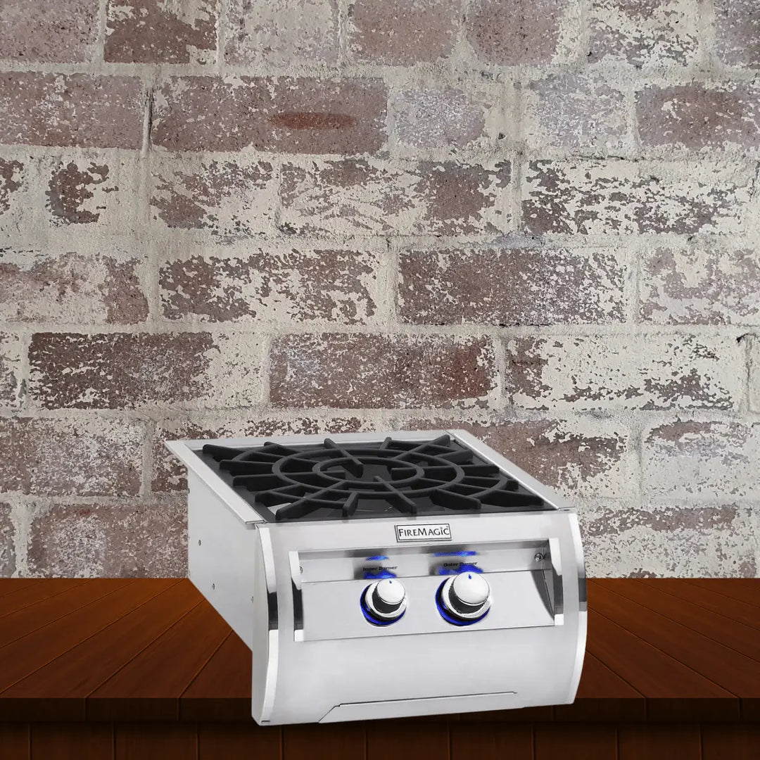 Fire Magic side cookers collection featuring a stainless steel outdoor side burner with dual control knobs and heavy-duty cast iron grate, displayed on a wooden surface with a rustic brick wall background.